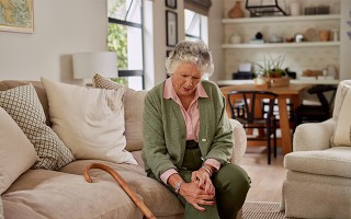 Ältere Frau sitzt auf dem Sofa und hält sich schmerzhaft das Knie.