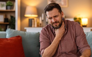 Mann sitzt auf dem Sofa und hält sich schmerzhaft den Hals, Hinweis auf Halsschmerzen.