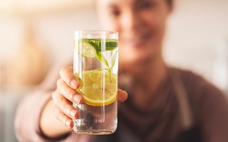 Eine Person hält ein erfrischendes Glas Wasser mit Zitronen- und Gurkenscheiben direkt in die Kamera.