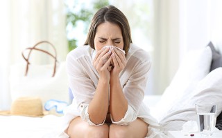 Junge Frau sitzt krank im Bett und putzt sich mit geschlossenen Augen die Nase.