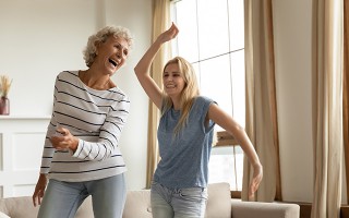 Ältere Frau und junge Frau tanzen fröhlich im Wohnzimmer.
