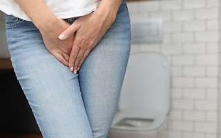 Frau hält Unterleib, Toilette im Hintergrund