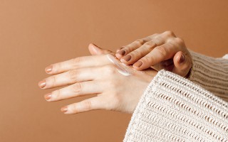 Person trägt eine weiße Handcreme auf den Handrücken auf; Nahaufnahme der Hände vor warmem, braunem Hintergrund.