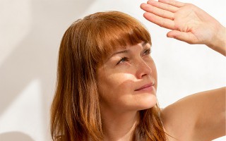 Eine rothaarige Frau blickt nach oben in die Sonne und schützt ihre Augen mit einer Hand vor dem hellen Licht.