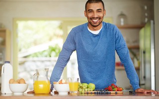 Mann steht lächelnd in der Küche vor frischem Obst, Saft und Milch.